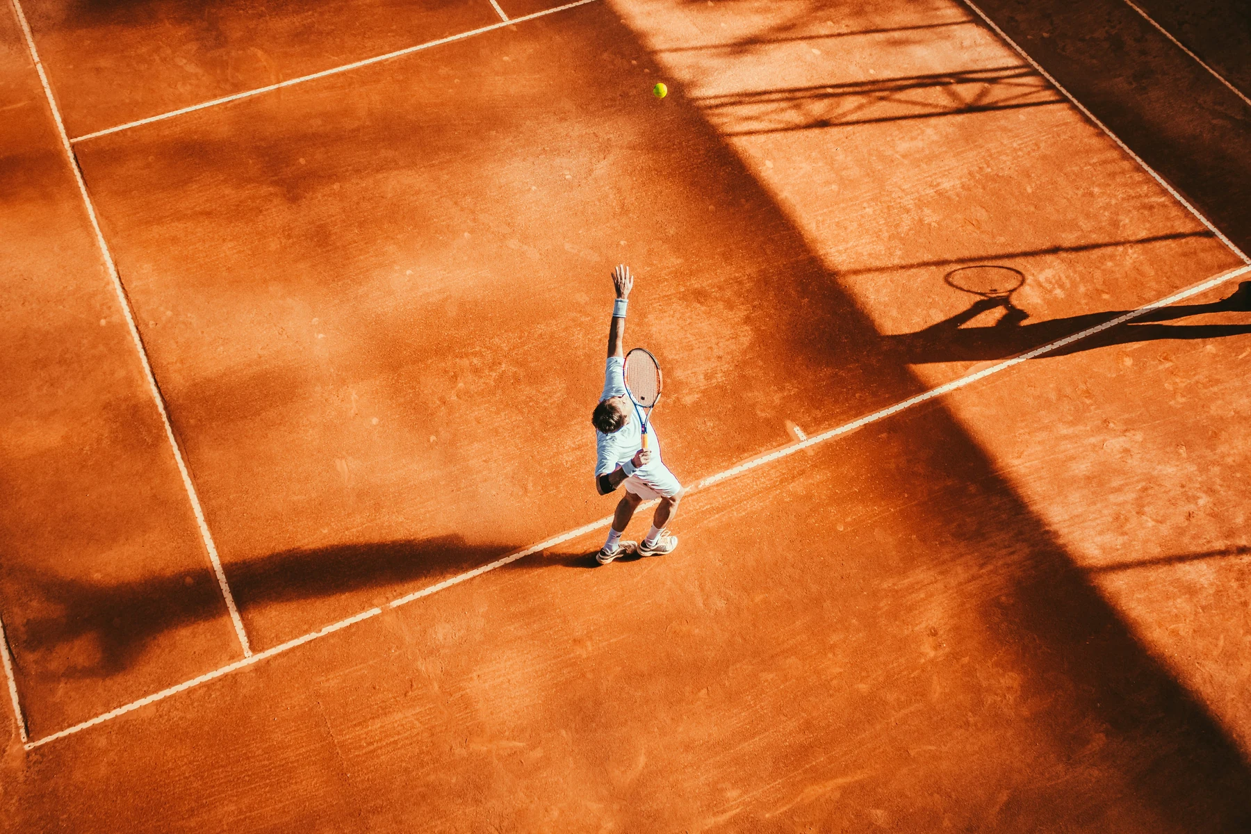 Overhead photograph of a tennis player tossing a ball for a serve on a sunlit clay court